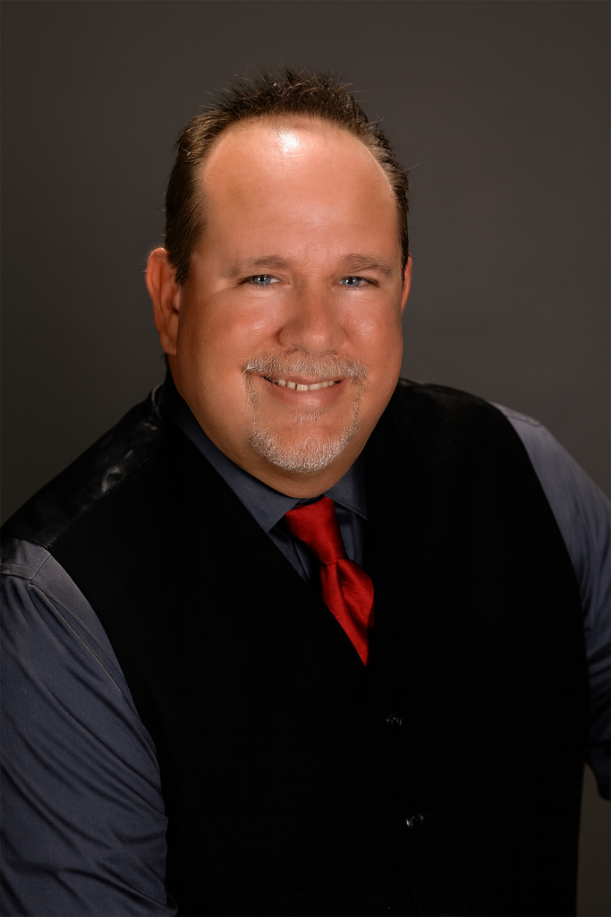 Professional author headshot of a smiling man wearing a black vest, gray shirt, and red tie against a studio background